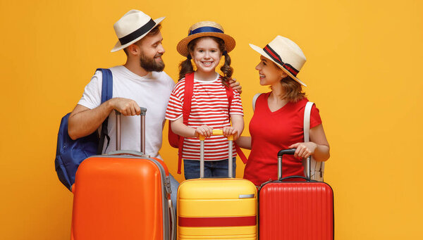 Optimistic parents and daughter leaning on luggage and smiling for camera during summer vacation against yellow backdro