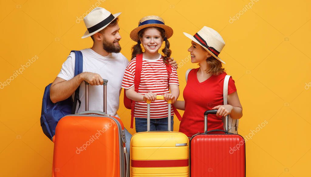 Optimistic parents and daughter leaning on luggage and smiling for camera during summer vacation against yellow backdro