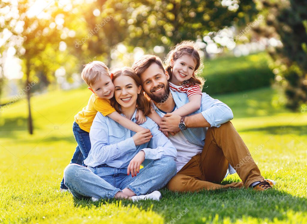 Cheerful boy and girl embracing happy parents from behind and looking at camera while resting on green grass on sunny day in par