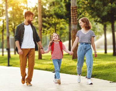 Tüm vücuduyla neşeli anne ve baba akıllı okul kızıyla el ele tutuşuyor ve yeşil yolda yürürken iletişim kuruyor.