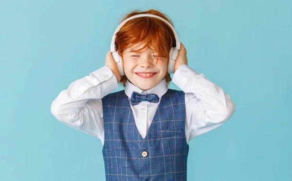 陽気生姜男の子で学校の制服目を閉じて笑顔で音楽を聴きながらヘッドフォンで青の背景 — ストック写真