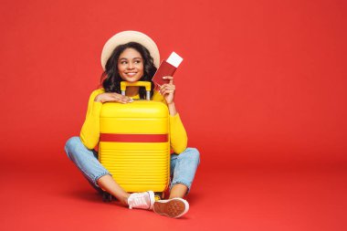 Full length positive ethnic tourist with passport and tickets smiling and dreaming about vacation while hugging suitcase against red backdro clipart