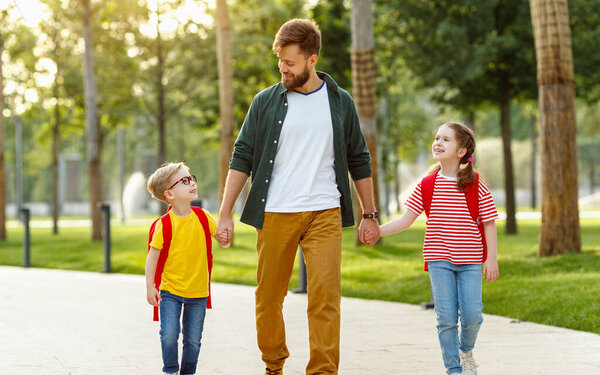 Bearded man smiling and holding hands with happy son and daughter while walking to school in park alle