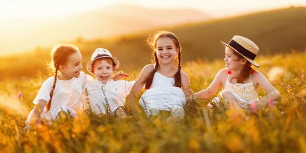 陽気な白い服を着た明るい小さな子供たちが草の中に座って 田舎で夏休みを楽しんでいる間に夕日のフィールドで一緒に楽しさと笑いを楽しんでいます — ストック写真