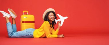 Şapkalı tam vücut etnik kadın valizin yanında yatıyor ve oyuncak uçakla Red Backdro 'ya karşı oynuyor.