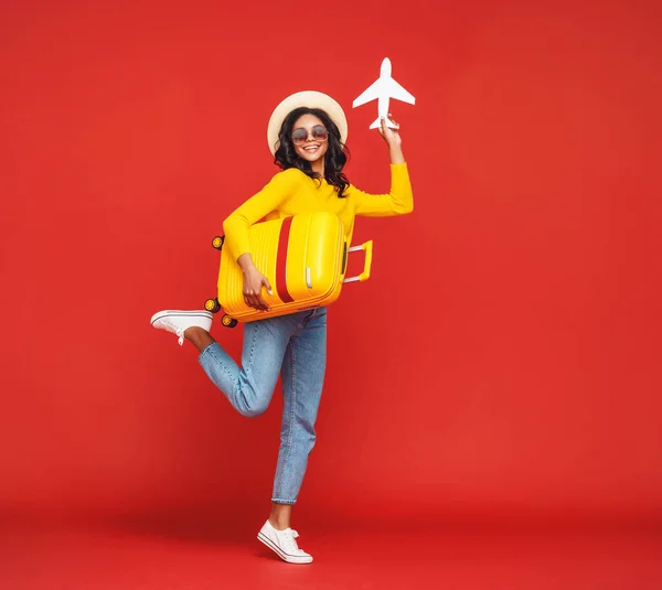 帽子とサングラスで全身民族の女性はスーツケースとジャンプし 赤い背もたれに対しておもちゃの飛行機で遊ぶ — ストック写真