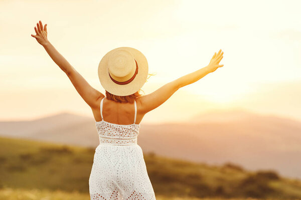 Обратный вид неузнаваемой молодой женщины в белом платье и шляпе, поднимающей руки и наслаждающейся свободой, стоя на зеленом лугу против размытых холмов и закатного неба во время летних каникул
