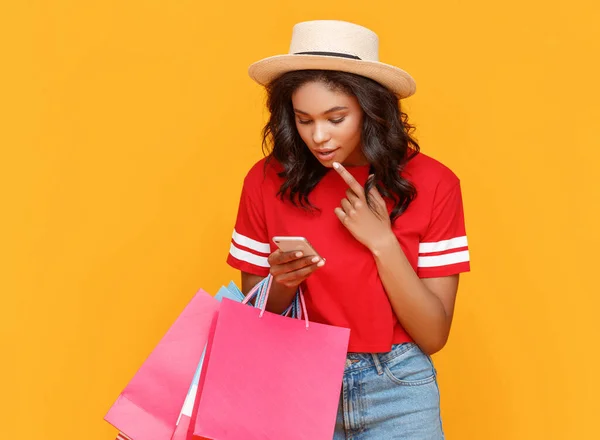 黄色の背景を背景にショッピング中に紙の袋を持つスタイリッシュな民族の女性は スマートフォン上であごとブラウジング割引オファーをピッキング — ストック写真