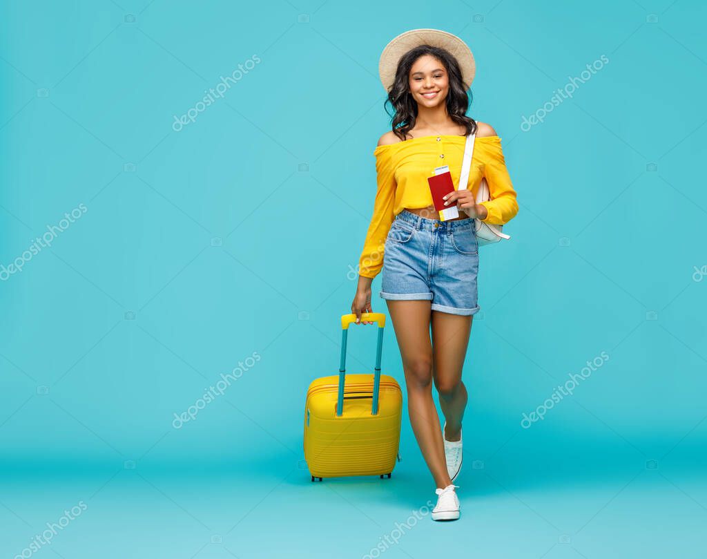 Full body positive ethnic woman with suitcase and tickets smiling and walking towards camera during trip against turquoise backdro