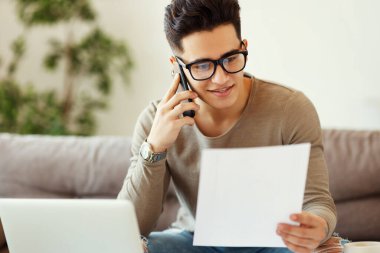 Mutlu etnik erkek, günlük giysiler içinde gülümseyip belgeleri okurken kanepede oturuyor ve evdeki telefonlara cevap veriyor.