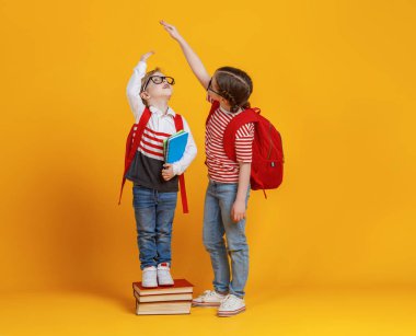 Küçük çocuk kitap yığınının üzerinde duruyor ve sarı sırt çantasıyla okula gitmeye hazırlanırken uzun boylu kıza ulaşmaya çalışıyor.
