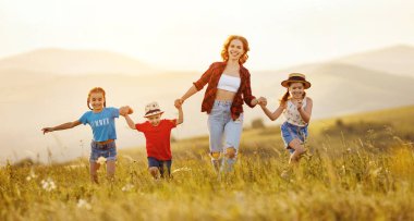 Sevimli küçük kızları ve oğlu olan neşeli genç bir kadın el ele tutuşup yeşil çayırlarda koşarken birlikte eğleniyorlar ve Natur 'da yaz akşamının tadını çıkarıyorlar.