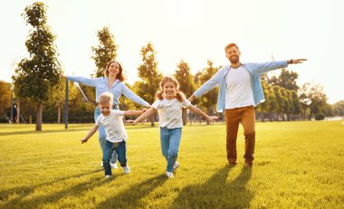 Heyecanlı ebeveynler ve çocuklar kollarını uzatmış gülümsüyor. Yaz parkındaki yeşil çimlerde kameraya doğru.