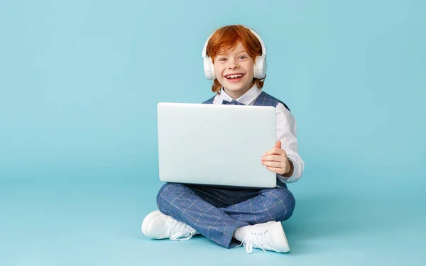 全身上下开朗的生姜男孩笑着 一边在笔记本电脑上做作业一边看着相机 双腿交叉坐在蓝色的背衬下 — 图库照片