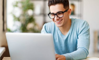 Evdeki masada ödev yaparken laptopta gülümseyen ve veri tarayan mutlu etnik genç adam.