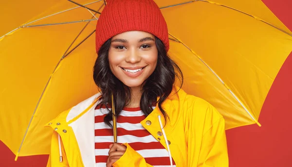 Positive ethnic female in hat and raincoat smiling and looking at camera while standing under umbrella on rainy day against red backgroun