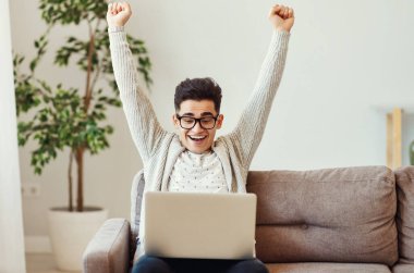 Kollarını kaldırmış, gülümseyen ve online piyangoda zaferi kutlayan genç etnik bir adam kanepede oturup dizüstü bilgisayar kullanıyor.