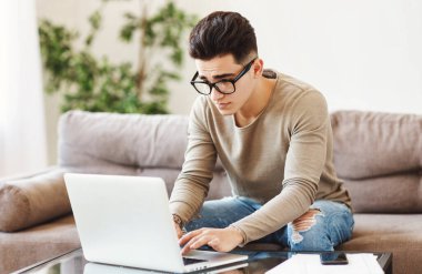 Sıradan giysiler ve gözlüklerle kanepede oturup evdeki uzak projeler üzerinde çalışırken laptopta veri taraması yapan genç etnik bir adam.