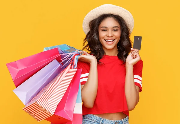 カラフルな紙袋の束を持つ喜びの民族の女性は笑顔と黄色の背景に対する買い物中にクレジットカードを示しています — ストック写真