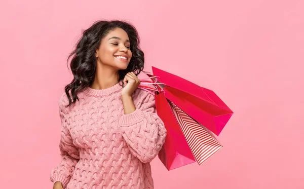 Positive ethnic woman smiling for camera and carrying paper bags on shoulder during shopping against pink backgroun