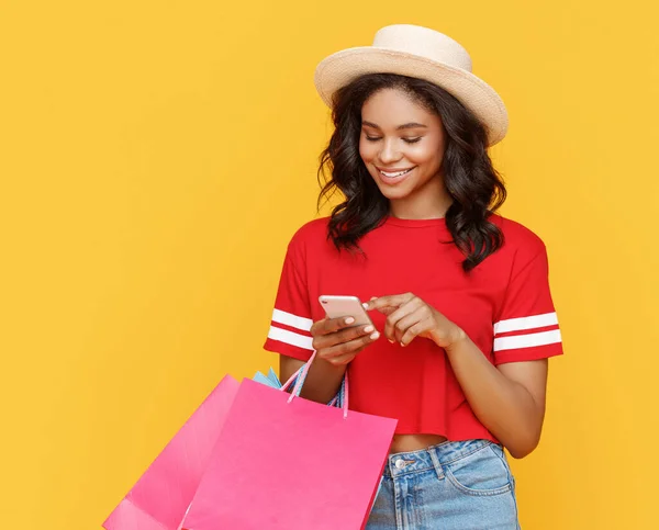 Happy ethnic woman with paper bags smiling   and browsing smartphone during shopping against yellow backgroun