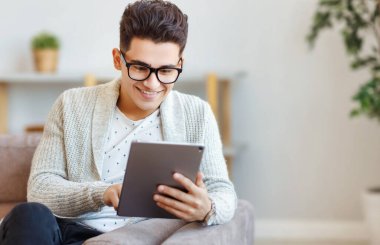 Evdeki rahat kanepede dinlenirken tablette gülümseyen ve sosyal medyada gezinen gözlüklü, mutlu etnik erkek.