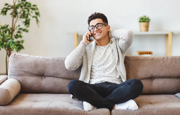 Full body happy ethnic man in casual clothes and glasses rubbing neck and looking away with smile while sitting cross legged on couch and answering phone call at hom