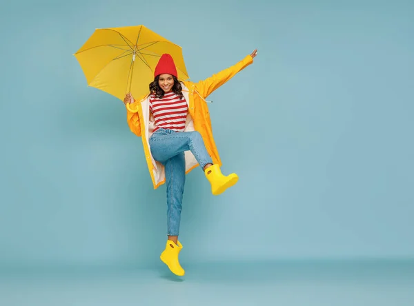 Full body happy ethnic woman in stylish outfit carrying umbrella and jumping on blue backgroun