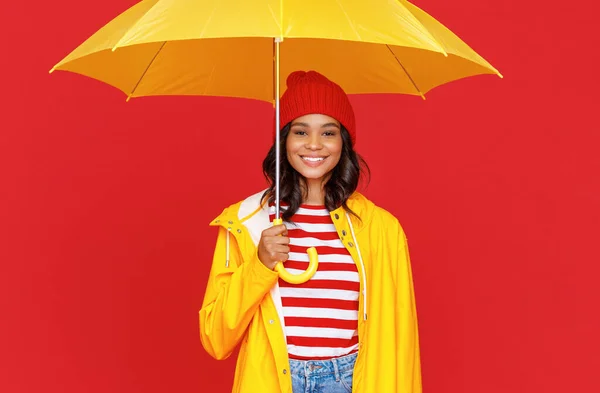 雨の日に傘の下に赤い背景を背に立ちながら 帽子とレインコートの笑顔とカメラを見て肯定的な民族女性 — ストック写真