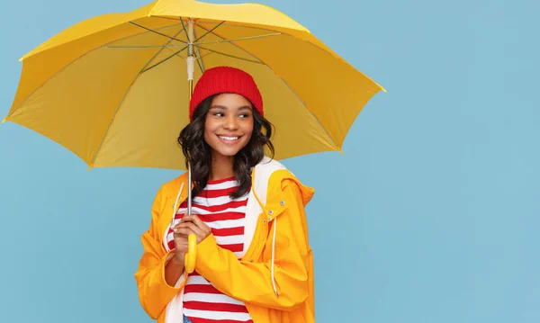 站在雨伞下 站在蓝色背衬下 戴着帽子 身穿雨衣的积极的少数民族女性笑了 — 图库照片