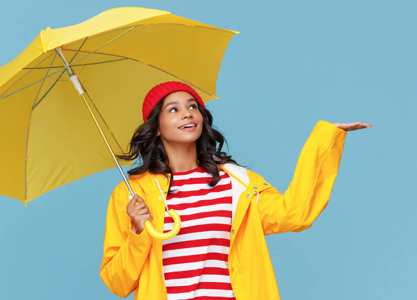 Cheerful ethnic female with yellow umbrella smiling and catching raindrops on rainy autumn day against blue backgroun