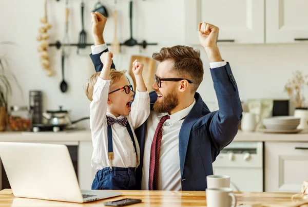 若いです男性起業家でフォーマルな服や眼鏡で小さな息子でフォーマルウェアを見てラップトップ画面とお祝い成功した契約ながら リモートで現代のアパートメンバーで作業 — ストック写真