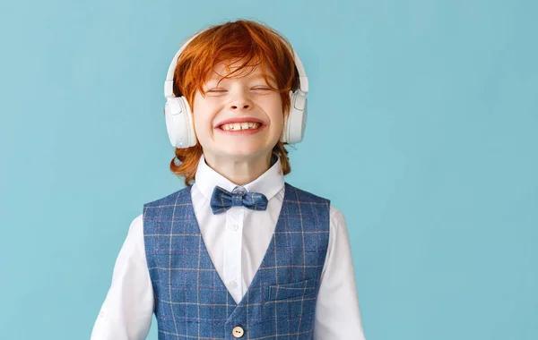 陽気生姜男の子で学校の制服で目を閉じて笑顔で音楽を聞きながらヘッドフォンで青の背景 — ストック写真