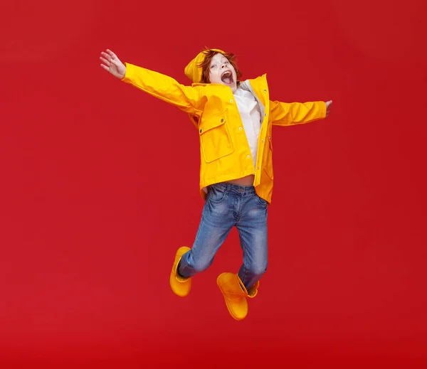 フルボディ幸せな男の子で黄色レインコートとブーツ大声でとLeapupでアウト伸ばさ腕ながら楽しいです雨の日に対して赤Backdro — ストック写真
