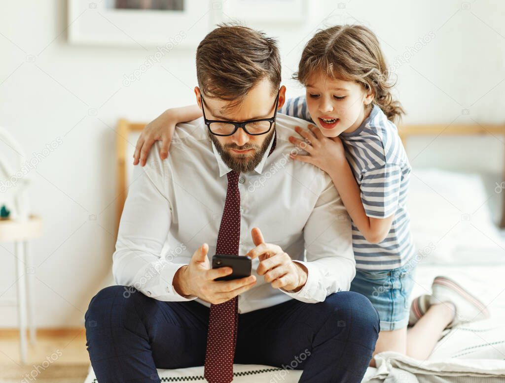 Serious frowned busy man in formal wear and eyeglasses reading business news on smartphone while cheerful little daughter sitting nearby and asking for attention during morning in bedroom at hom