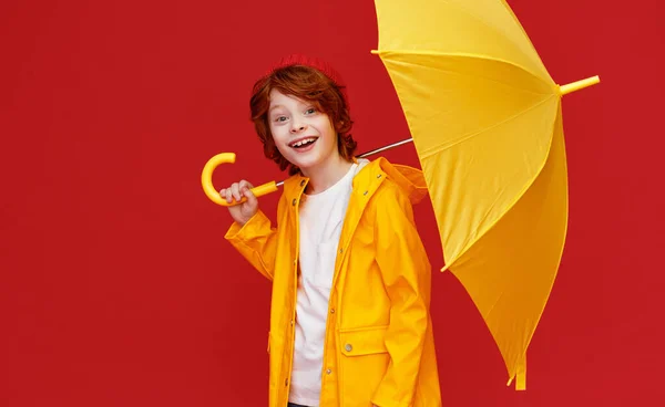 陽気男の子でレインコート笑顔のためカメラと運ぶ黄色傘肩に雨の日に対して赤 — ストック写真
