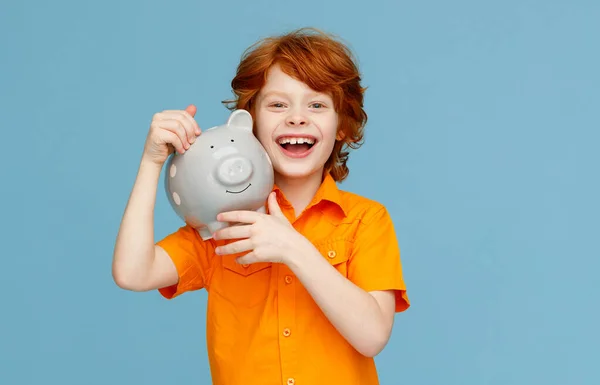 快乐的红头发男孩 身穿橙色衬衫 高兴地对着相机笑着 肩并肩抱着小猪 背靠着蓝色的背脊 — 图库照片