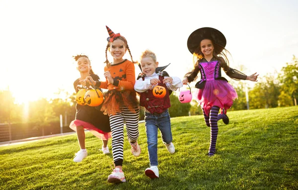 Group of excited kids in spooky costumes smiling and running on lawn during Halloween celebration in evening in par