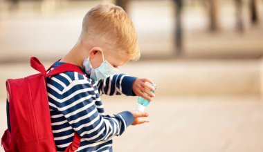 Korunaklı maskeli küçük ilkokul çocuğu Coronavirus pandemi sırasında antibakteriyel jelle ellerini temizlerken.