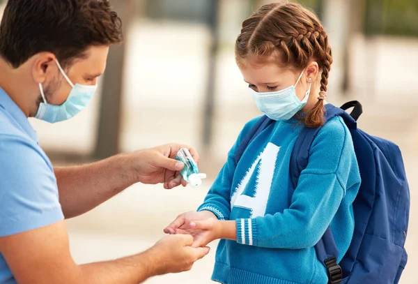 Side view of careful dad in protective mask pouring sanitizer into hands of little school aged daughter during coronavirus pandemi