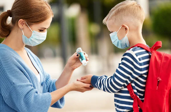 Side view of careful mom in protective mask pouring sanitizer into hands of little school aged son during coronavirus pandemi