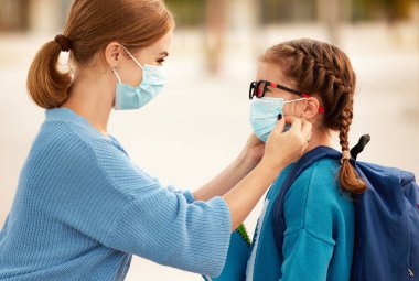 Tanınmayan anne, Coronavirus pandemi sırasında okula hazırlanırken neşeli okul kızının yüzüne tıbbi maske takıyor.