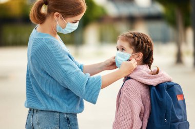 Tanınmayan anne, Coronavirus pandemi sırasında okula hazırlanırken neşeli okul kızının yüzüne tıbbi maske takıyor.