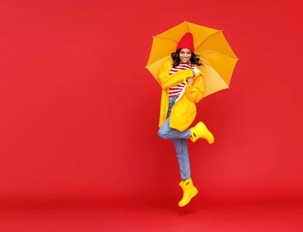 Full body delighted ethnic woman in raincoat smiling and leaping up on rainy autumn day against red backgroun