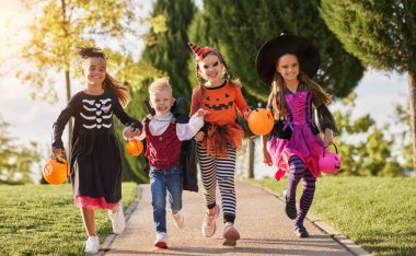 Group of excited kids in spooky costumes smiling and running on path in  during Halloween celebration in evening in par clipart