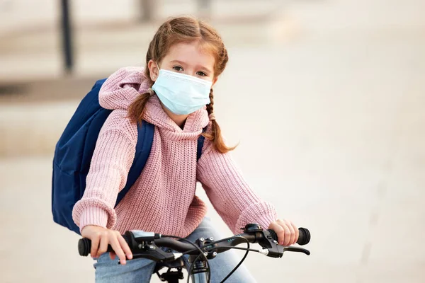 预防眼镜蛇 19和病毒感染的概念 穿着保暖针织服装 戴着防护罩 背着背包 骑着自行车在城市里骑车时看着相机的积极女学生 — 图库照片