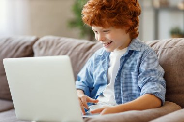 Evdeki yumuşak kanepede otururken gündelik giysiler içinde gülümseyen ve laptopta oyun oynayan neşeli kızıl çocuk.