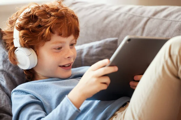 穿着休闲装和耳机的红头发男孩快乐的坐在沙发上玩平板电脑游戏 — 图库照片