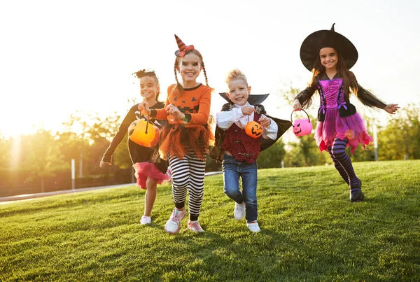 Group of excited kids in spooky costumes smiling and running on lawn during Halloween celebration in evening in par