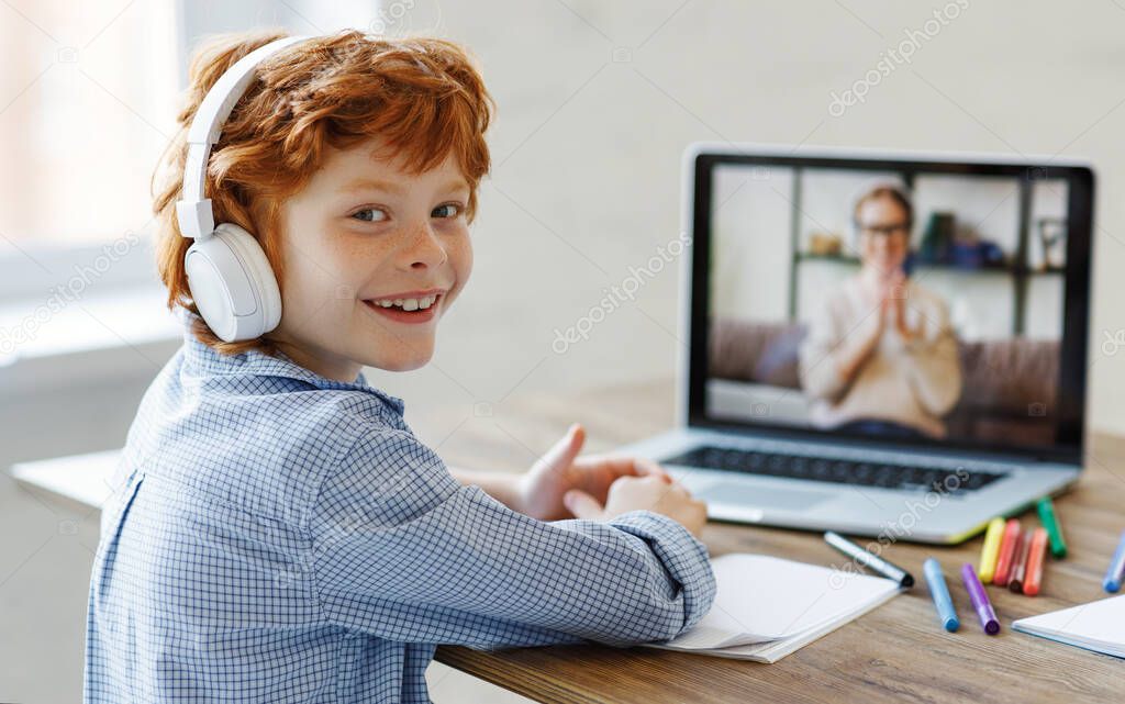 Happy boy in headphones smiling and looking at camera while making video call to teacher during online lesson at hom